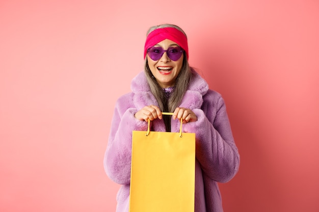 Fashionable asian senior woman going shopping in sunglasses and trendy winter clothes, holding paper bag from store and smiling happy at camera, pink background.