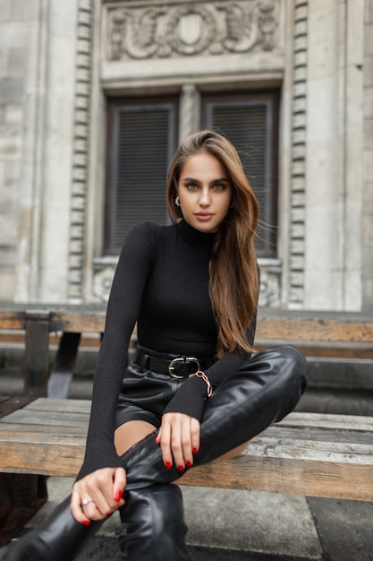 Fashionable beautiful elegant woman vogue model in stylish black clothes with a long sleeve shirt leather shorts and high boots sits on a wooden bench near a vintage building in a European city