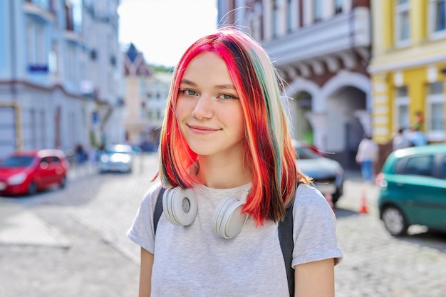 Fashionable beautiful girl teenager 16, 17 years old in wireless headphones with bright dyed colored hairstyle on the street of summer sunny city looking at camera. Lifestyle, youth, fashion, beauty