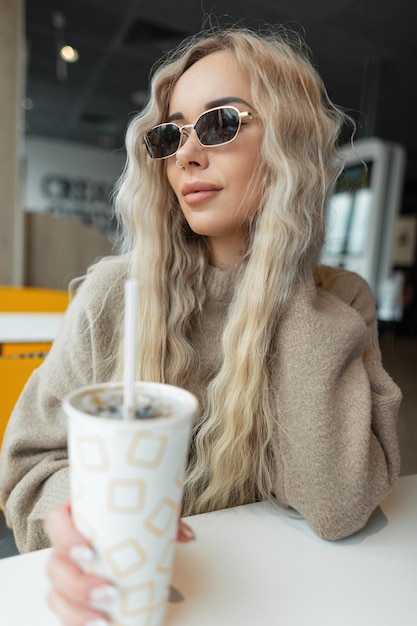 Fashionable beautiful girl with a curly blond hairstyle with cool su sunglasses in a beige sweater holds a paper cup of cola and sits in a cafe