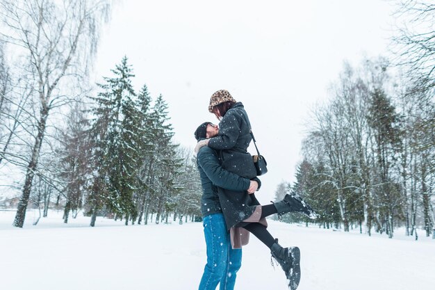 Fashionable beautiful happy couple of lovers in fashion winter clothes having fun in a winter park with snow Handsome man picked up his girlfriend woman