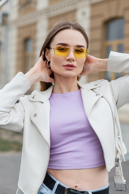 Fashionable beautiful hipster street girl with stylish yellow sunglasses and purple Tshirt with leather jacket in the city