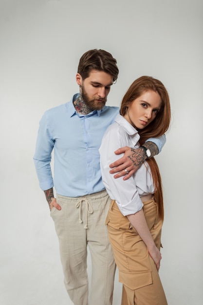 Fashionable beautiful lovers couple pretty redheaded girl and hipster guy with a tattoo and beard in stylish clothes with a shirt on a white background in the studio