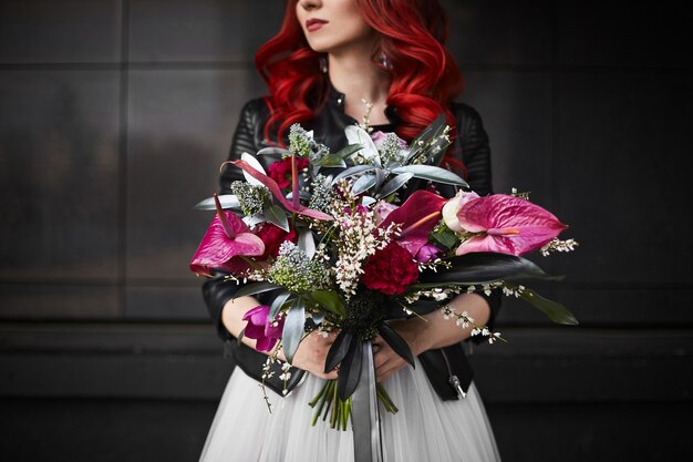 Photo fashionable and beautiful model girl with red hair and bright makeup, in a white wedding dress and in a leather jacket, with a big and luxury bouquet of exotic flowers in her hands, posing outdoors