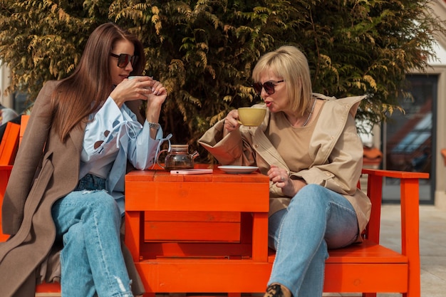 Fashionable beautiful mother and daughter in stylish casual clothes sit in a cafe on the street and drink tea and coffee on a spring sunny day