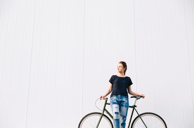 Fashionable beautiful woman standing with bicycle, looking away, over white wall. Copy Space. Outdoors