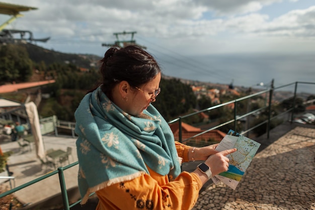 Fashionable beautiful woman with sunglasses in a stylish yellow dress with a vintage green scarf holds a map and travels on the island of Madeira Portugal