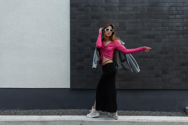 Fashionable beautiful young redheaded hipster woman with cool sunglasses wearing a trendy pink top with a long skirt sneakers and jeans jacket walks near a black brick wall on the street