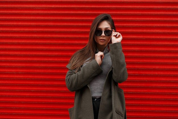 Fashionable beautiful young woman in a  fashion coat and gray sweater straightens stylish sunglasses near the red metal wall