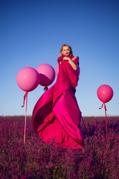 Fashionable beautiful young woman on a pink stepladder in a pink dress in a field with wildflowers in summer at sunset