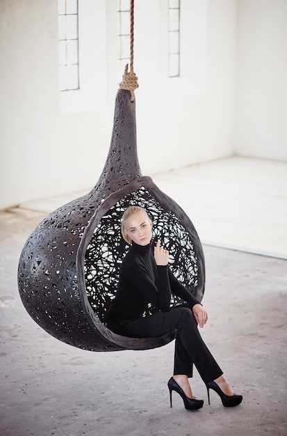 Fashionable blond female sits on a round chair in a big empty room.