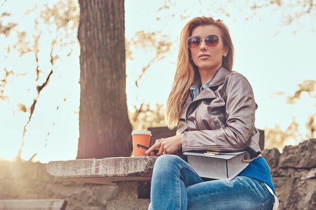 Photo fashionable blonde blogger female relaxes outdoor, using a smartphone while sitting on the bench in city park against a bright sunlight.