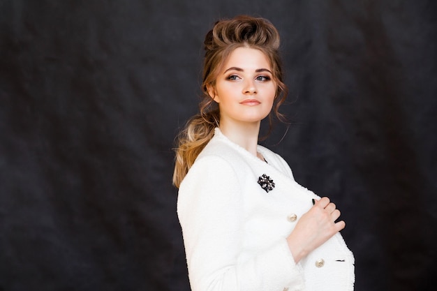 Fashionable brunette girl posing on a black background in a stylish white jacket with a brooch in the form of a flower rhombus