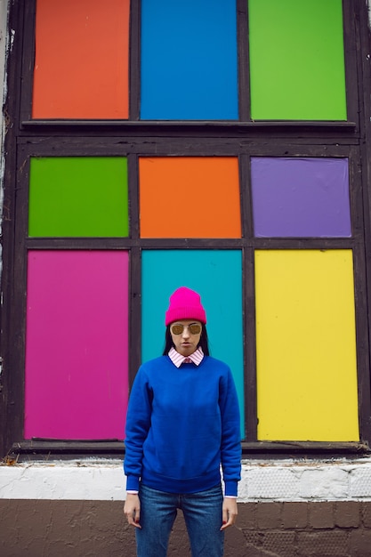 Photo fashionable brunette woman in a pink warm hat and sweater and sunglasses stands against the background of colorful windows in autumn