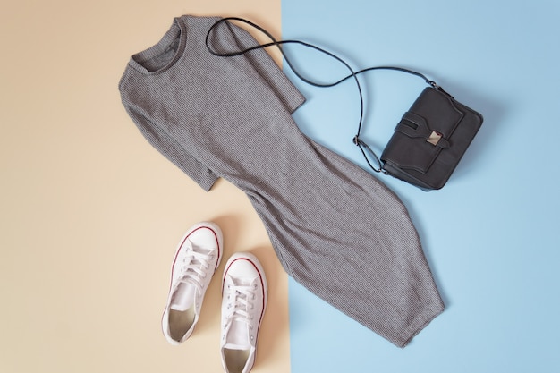 Fashionable concept. Women's urban style. Gray dress, white sneakers and black handbag on a soft blue background, beige