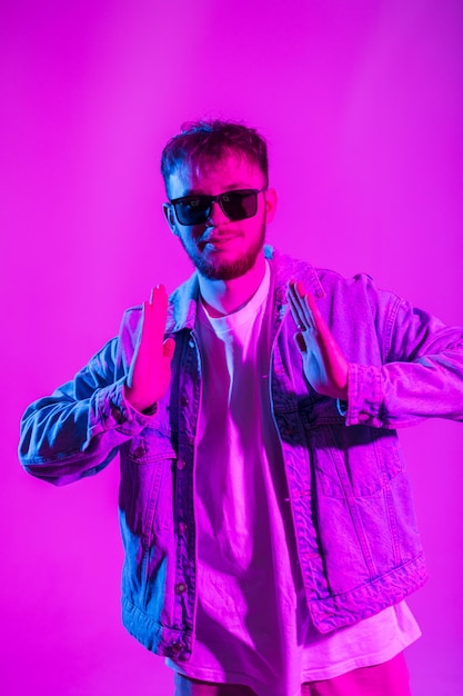 Fashionable cool dancer guy with sunglasses in denim jacket dances in creative colorful studio with pink and blue light