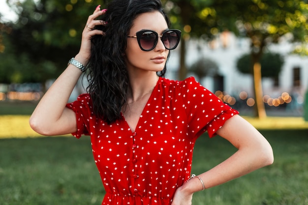 Fashionable curly woman with sunglasses in vintage red dress posing on the street