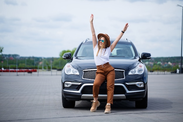 Fashionable image of a stylish young girl in a hat and white T-shirt. A girl stands near a black car with a smile on her face