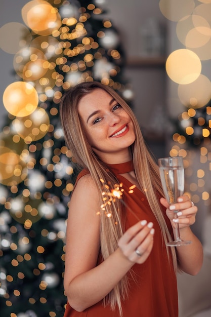 Fashionable interior beautiful woman in a blouse posing near a decorated Christmas tree holding champagne and a sparkler