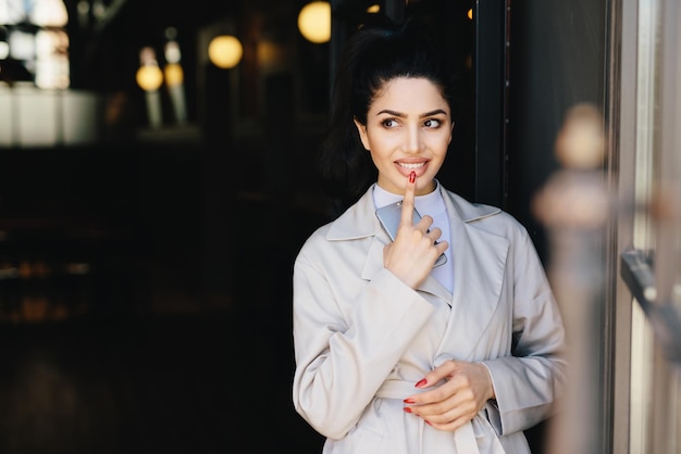 Fashionable lady with dark luminous hair tied in pony tail having nice eyes and red manicure holding her indexfinger on full lips and cell phone in hand having mysterious expression looking aside