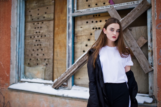 Fashionable long legs brunette model in long black cloak posed outdoor at winter day against old grunge wall with broken windows.