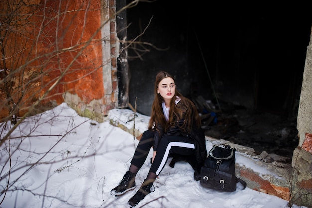 Fashionable long legs brunette model in long black cloak posed outdoor at winter day against old grunge wall.