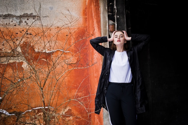 Fashionable long legs brunette model in long black cloak posed outdoor at winter day against old grunge wall.