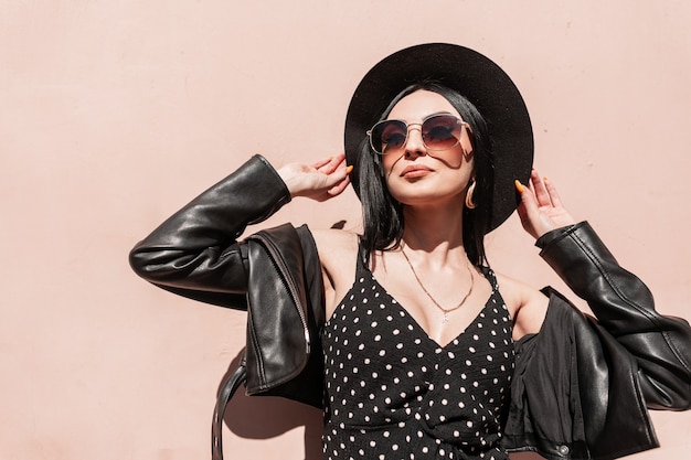 Fashionable lovely woman in hat in jacket in dress in sunglasses enjoys sun in city. Attractive fashion girl model in black elegant summer beautiful clothes posing in bright sunlight near pink wall