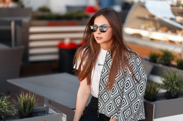 Fashionable model girl in sunglasses and fashionable jacket sits in a cafe on the street