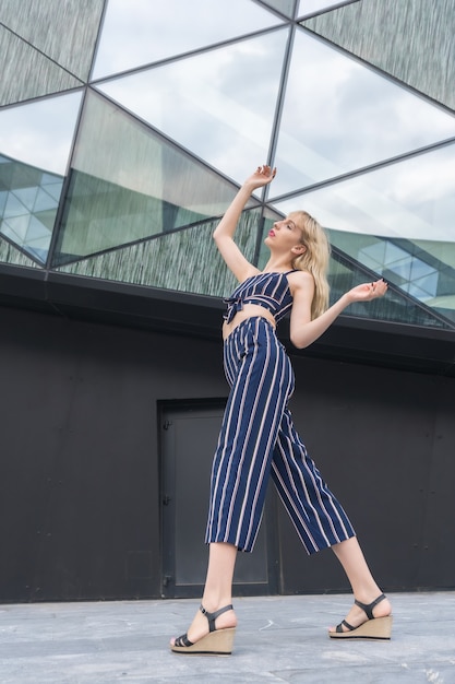 Fashionable posing of a young blonde in a glass building, blonde girl in a blue striped suit, arms up