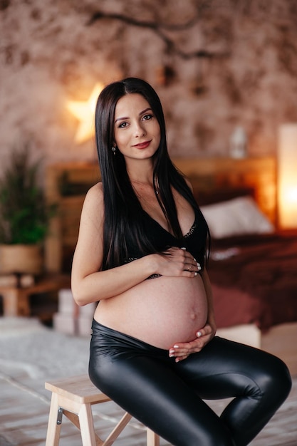 Fashionable pregnant mom hugging belly Stylish Motherhood concept Happy young pregnant woman in stylish black bra and modern earring holding belly bump and posing in light fashion photo