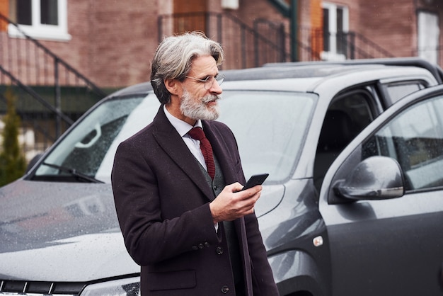 Fashionable senior man with gray hair and beard standing outdoors on the street near his car with smartphone in hand.