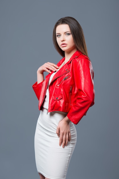 Fashionable style, fashion women's clothing, color combination. Beautiful brunette girl in white dress and red leather jacket isolated gray background.