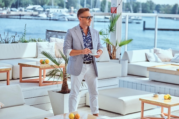 Photo fashionable successful man with stylish hair dressed in modern elegant clothes holds cup of coffee while standing at outdoor restaurant against the background of the city wharf.