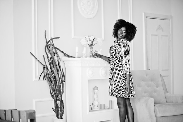 Fashionable tall african american model woman with red afro hair in dress posed at white room against yellow chair and fireplace