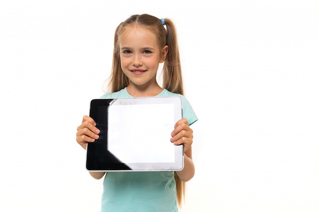 Fashionable teenager girl in blue t-shirt with tablet gesticulates and smiles on white wall