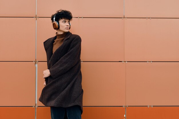 Fashionable woman in coat and scarf standing in front of orange wall with headphones listening to