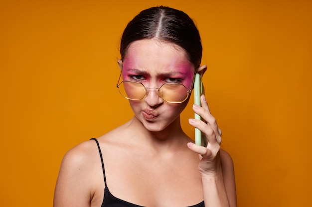 Fashionable woman with phone pink face makeup posing attractive look glasses yellow background unaltered