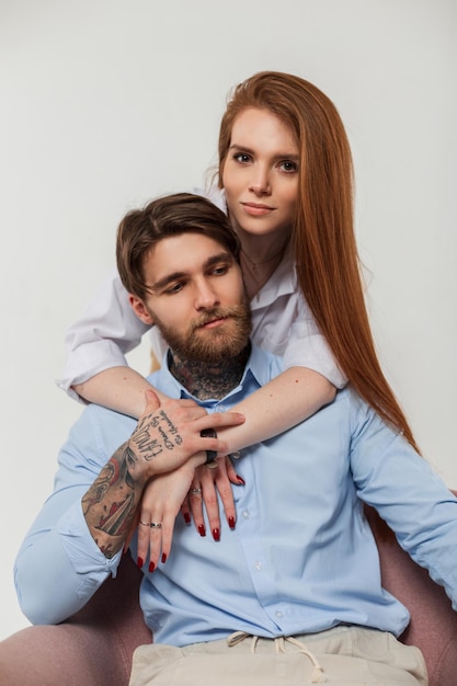 Fashionable young beautiful couple Pretty fashion redhead elegant woman hug a handsome hipster man with tattoos on a white studio