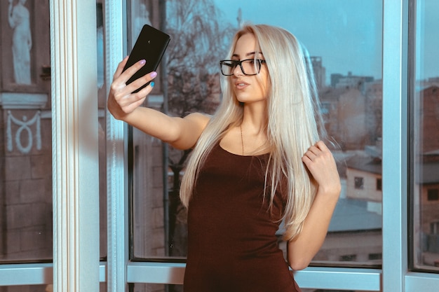 Fashionable young blonde lady makes selfie by the window with city views. Business woman makes selfie. City views behind the window in the office. Business concept.