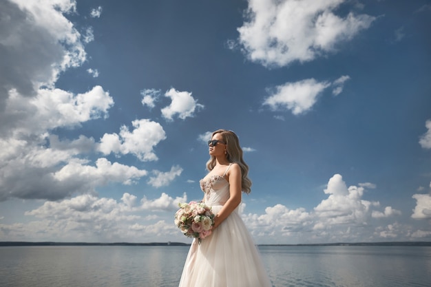 Fashionable young bride with platinum blond hair in wedding dress posing with bouquet of fresh flowers on the sea coast.