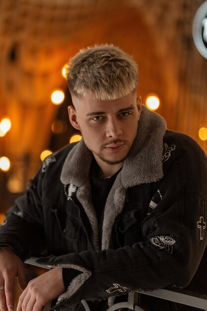 Fashionable young handsome man with hairstyle in trendy clothes with stylish jacket stands in the mall with light