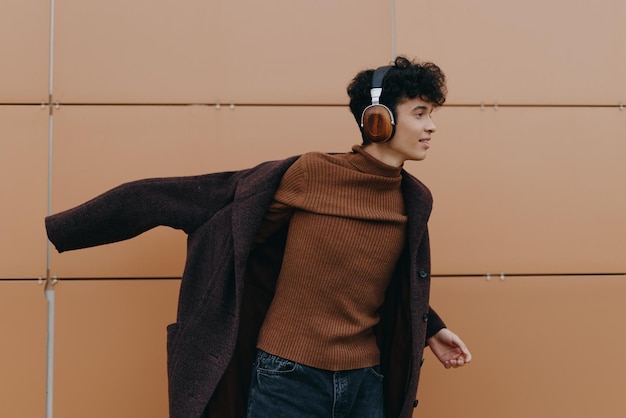 Fashionable young man in brown sweater and headphones smiling with arms outstretched against orange