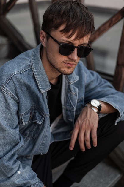 Fashionable young man hipster in stylish denim clothes trendy in a black sunglasses sits on a vintage staircase in the city on a spring day. Attractive cool guy resting outdoors. Trendy menswear.