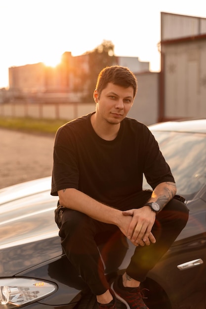 Fashionable young man model in stylish black summer clothes sits on the car at sunset