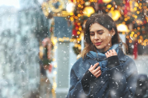 Fashionable young woman dressed in trendy coat walking at the street during the snowfall. Empty space