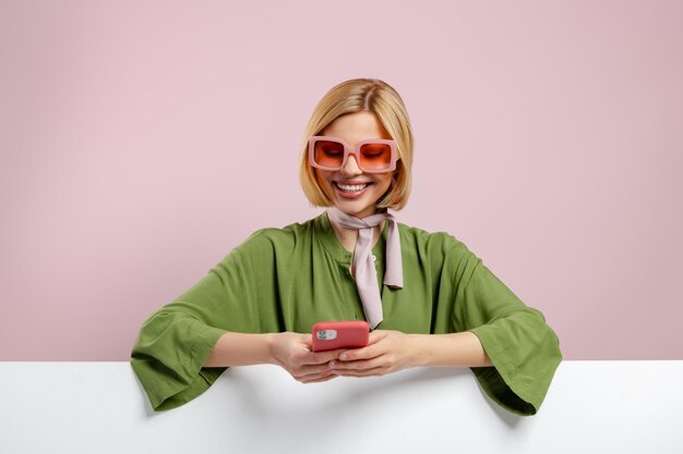 Photo fashionable young woman leaning at copy space and using phone against pink background