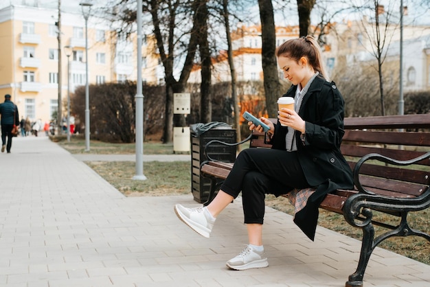 Fashionable young woman in stylish coat chatting on internet on mobile phone while sitting on park bench Side view of pretty lady holding smartphone and cup of coffee outdoors People and technology