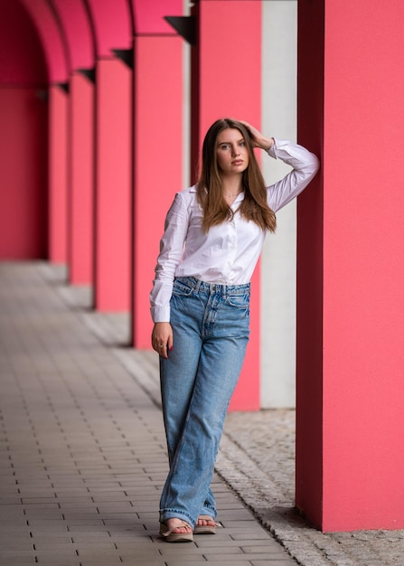 A fashionable young woman with long hair was leaning on a column Looks at the camera A Caucasian girl in a casual outfit walks through the city among pink columns