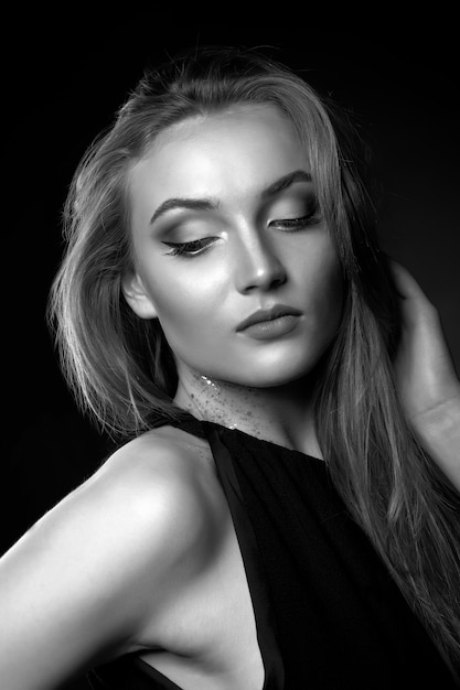 Fashionable young woman with long straight hair wears classic black dress posing in the shadow at studio. Black and white shot